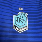 camiseta el barrio - kings league - escudo Primer plano del escudo del equipo 'El Barrio' de la King League en la camiseta oficial, sobre fondo azul con líneas horizontales, y detalle de dos coronas doradas encima del escudo.
