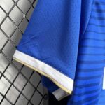 camiseta el barrio - kings league - manga Detalle de la manga de la camiseta 'El Barrio' de la King League en color azul, con borde en blanco y dorado, mostrando la textura perforada del material para mayor transpirabilidad.