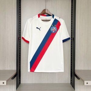 Descubre la camiseta Bahia primera equipación 2026-2027, una prenda que combina la tradición del fútbol brasileño con un diseño moderno y lleno de personalidad. Su base blanca contrasta con una llamativa banda diagonal en azul marino y rojo, creando una estética distintiva para los aficionados del Esporte Clube Bahia. Esta camiseta destaca por la disposición diagonal de sus colores, el escudo del club en el pecho y los detalles en rojo y azul que decoran las mangas y los hombros. El cuello redondo blanco mantiene una apariencia limpia y deportiva, mientras que la combinación cromática aporta un estilo reconocible para quienes siguen al conjunto de Salvador de Bahía. Un diseño blanco con la identidad del Bahia La primera equipación del Bahia apuesta por una composición visual diferente a las tradicionales camisetas de franjas verticales. El blanco domina la prenda y sirve de fondo para una banda diagonal que combina azul marino y rojo, dos tonos que destacan sobre el tejido. El escudo del Esporte Clube Bahia ocupa una posición protagonista en el pecho, acompañado por dos estrellas doradas. Los acabados de las mangas alternan rojo y azul, reforzando la personalidad del diseño y creando una camiseta atractiva tanto para los partidos como para el uso diario. Si buscas una camiseta del Bahia 2026-2027 con un estilo diferente, esta primera equipación es una opción interesante para incorporar los colores del fútbol brasileño a tu colección. Comodidad deportiva para jugar y entrenar Además de su diseño, esta camiseta está pensada para acompañarte en distintas actividades deportivas. Su construcción ligera y su corte deportivo favorecen la comodidad durante el movimiento, por lo que puedes utilizarla para jugar partidos con amigos, entrenar o practicar fútbol de manera habitual. Su silueta deportiva permite combinarla con pantalones cortos de fútbol y otras prendas de entrenamiento. También puedes llevarla en tus actividades cotidianas para mostrar tu afición por el Bahia y por el fútbol sudamericano. El orgullo de vestir los colores del Esporte Clube Bahia El Esporte Clube Bahia es uno de los clubes históricos de Salvador, en el estado brasileño de Bahía. Sus seguidores mantienen una fuerte vinculación con los colores y la identidad del equipo, que cuenta con una trayectoria destacada dentro del fútbol nacional. Esta primera equipación representa una alternativa para quienes quieren seguir al Bahia, descubrir camisetas de clubes brasileños o ampliar una colección de equipaciones internacionales. Su base blanca y su banda diagonal roja y azul aportan una imagen diferente a la de muchas camisetas de fútbol convencionales. Características de la camiseta Bahia 2026-2027 Equipo: Esporte Clube Bahia. Temporada: 2026-2027. Tipo: primera equipación. Color principal: blanco. Diseño frontal: banda diagonal azul marino y roja. Cuello: redondo blanco. Mangas: detalles rojos y azules en los puños. Hombros: detalles de color. Escudo: Esporte Clube Bahia en el pecho. Estilo: deportivo y moderno. Uso recomendado: fútbol, entrenamientos y actividades cotidianas. Ideal para: aficionados del Bahia y coleccionistas de camisetas de fútbol brasileñas. ¿Por qué elegir la camiseta Bahia primera equipación? La camiseta Bahia 2026-2027 reúne una estética deportiva y una combinación de colores muy reconocible. Su banda diagonal roja y azul sobre el fondo blanco le proporciona una personalidad propia, mientras que los detalles de las mangas y el escudo completan el diseño. Si buscas una camiseta del Esporte Clube Bahia para jugar, entrenar o vestir los colores de un equipo histórico de Brasil, esta primera equipación es una excelente opción para tu colección de ropa futbolística.